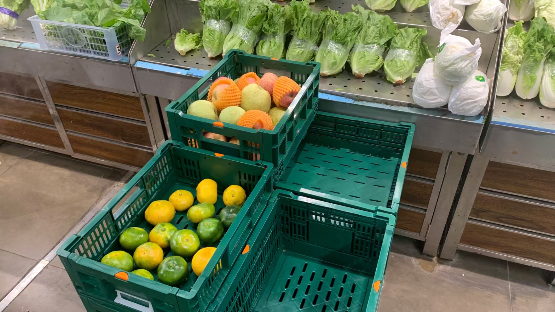 Cajas de plástico plegables de color verde utilizadas para exponer fruta en un supermercado, con diseño apilable y ventilado
