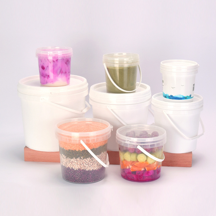 White opaque and transparent PP round plastic buckets in two sizes displayed together, small transparent buckets filled with colorful food contents including fruit drinks, smoothies, mixed grains, and candy, arranged on wooden display stand with light grey background